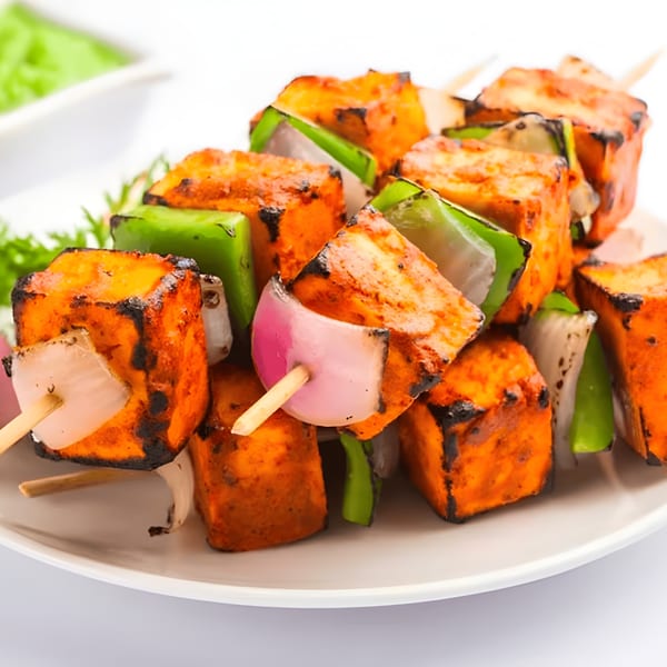 Grilled paneer tikka skewers with bell peppers and onions, served on a white plate with green chutney.